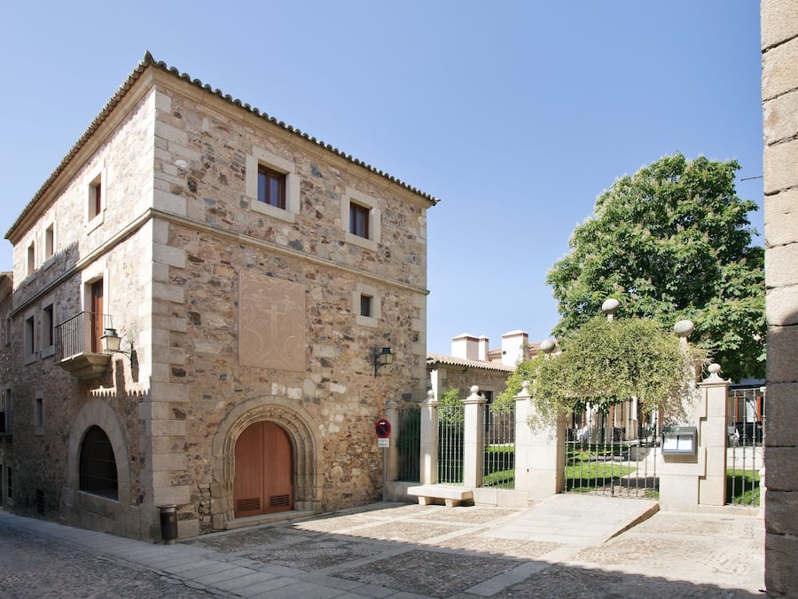 Un edificio de piedra con puerta de madera y ventanas arqueadas, rodeado por un patio empedrado y un árbol.