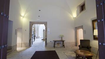 A hallway with a carpeted floor, a wooden table with a vase, a patterned armchair, and a mirror on the wall.
