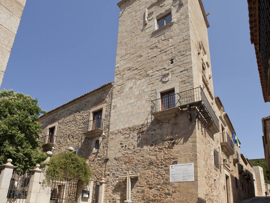 Um edifício histórico de pedra com uma torre alta, entrada com portão e pátio pavimentado.
