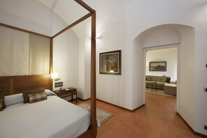 A bedroom with a four-poster bed, a nightstand with a lamp, a painting on the wall, and a view into a living area with a sofa and a table. - Parador de Cáceres (Cáceres)
