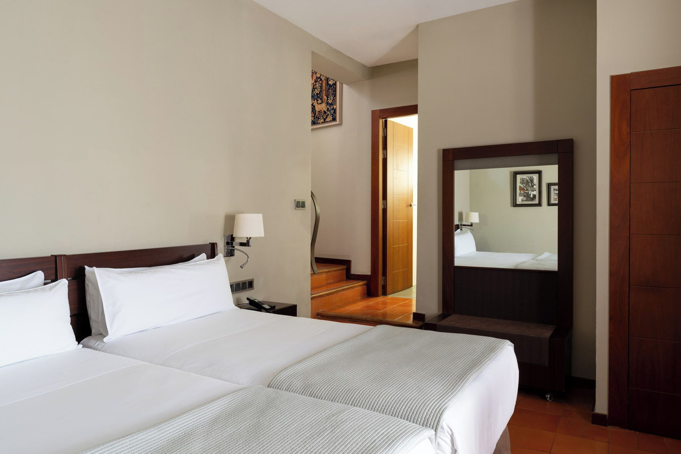 Ein Hotelzimmer mit zwei Betten, einem Nachttisch, einer Lampe, einem Spiegel und einer Holztür.