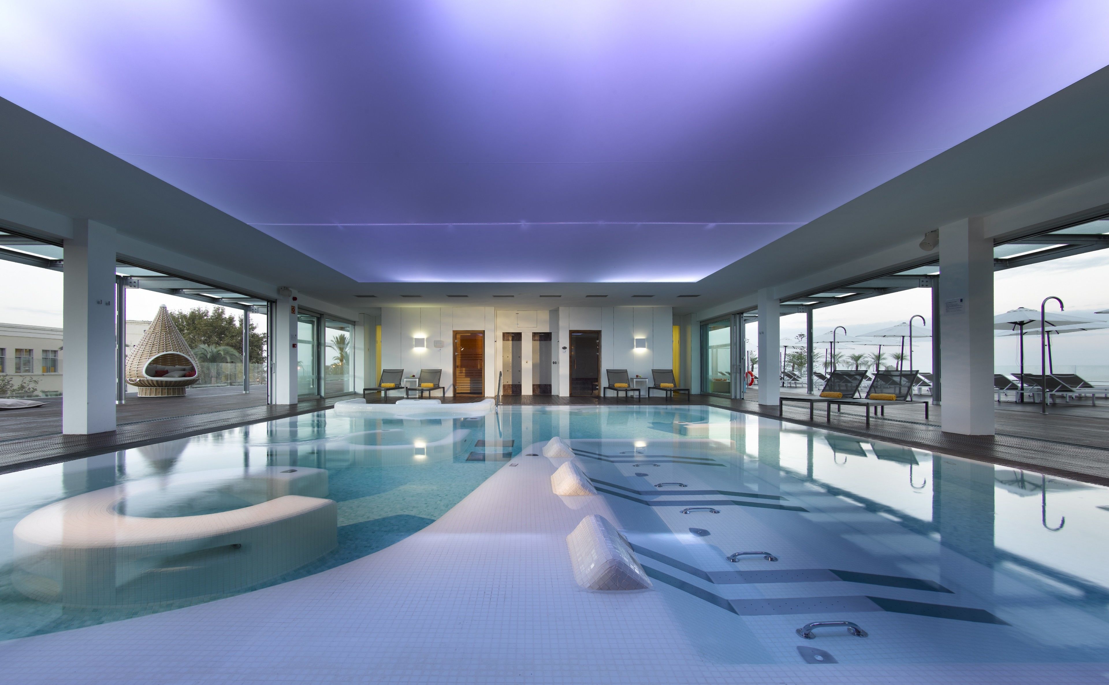 A large indoor swimming pool with clear water, surrounded by lounge chairs and a modern architectural design.