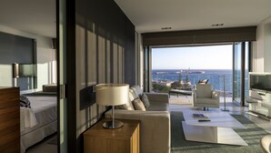 Una habitación de hotel moderna con una cama grande, un sofá, una mesa de centro y un televisor. La habitación tiene un ventanal con vistas al mar y al paisaje urbano.