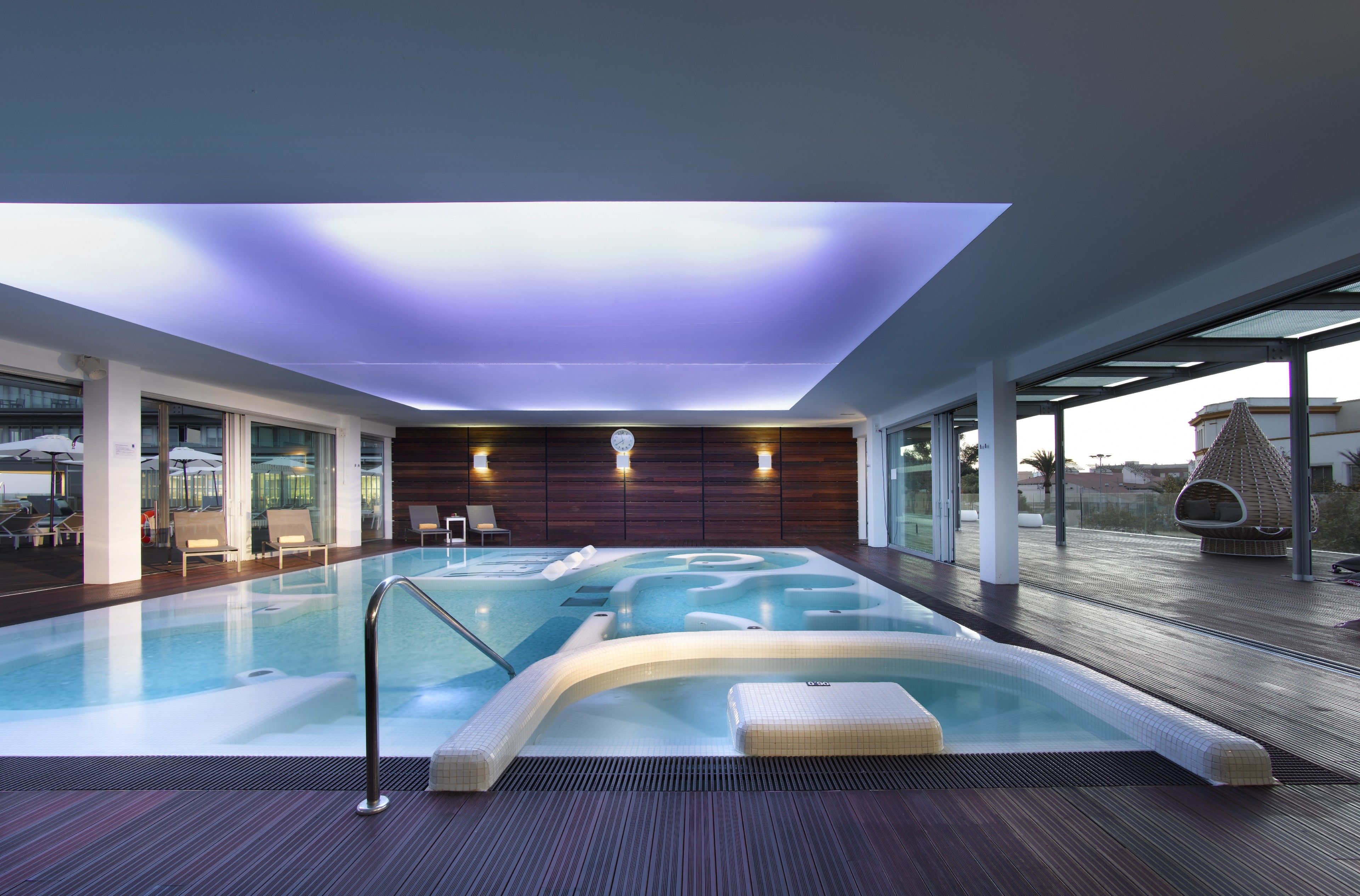 Une piscine intérieure moderne dotée d’un toit en verre, d’une terrasse en bois et de chaises longues.