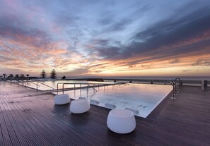 A wooden deck with a pool and seating area, overlooking a body of water at sunset. - Parador De Cadiz (Cádiz)