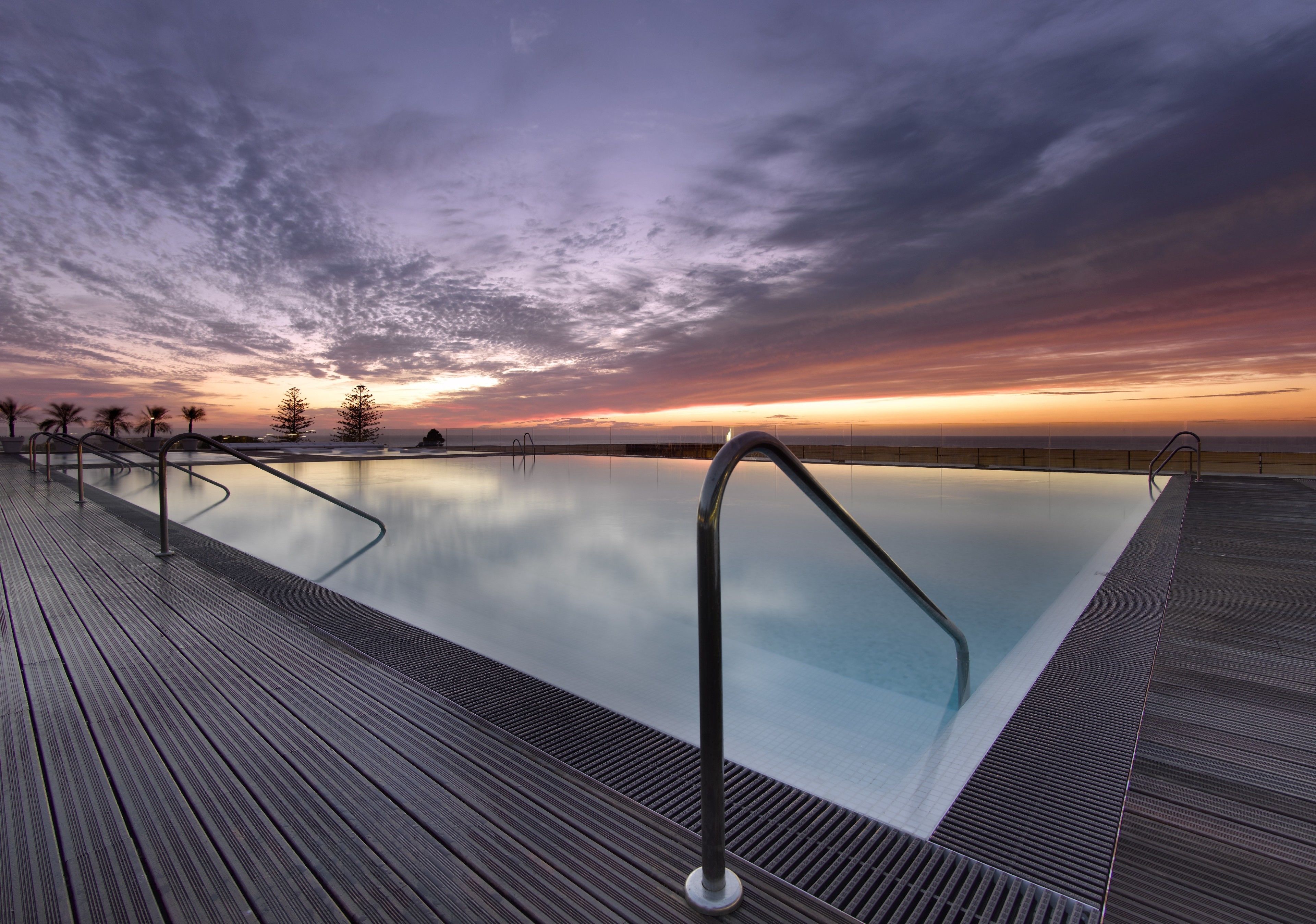 Una piscina infinita con cubierta de madera y barandillas de metal, frente a un cielo al atardecer.