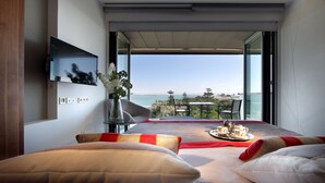 Una habitación de hotel moderna con una cama grande, un televisor de pantalla plana montado en la pared, una mesa con un servicio de desayuno y un balcón con vistas al mar.