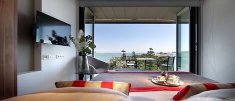 A modern hotel room with a large bed, a flat-screen TV mounted on the wall, a table with a tray of breakfast items, and a balcony with a view of the sea.