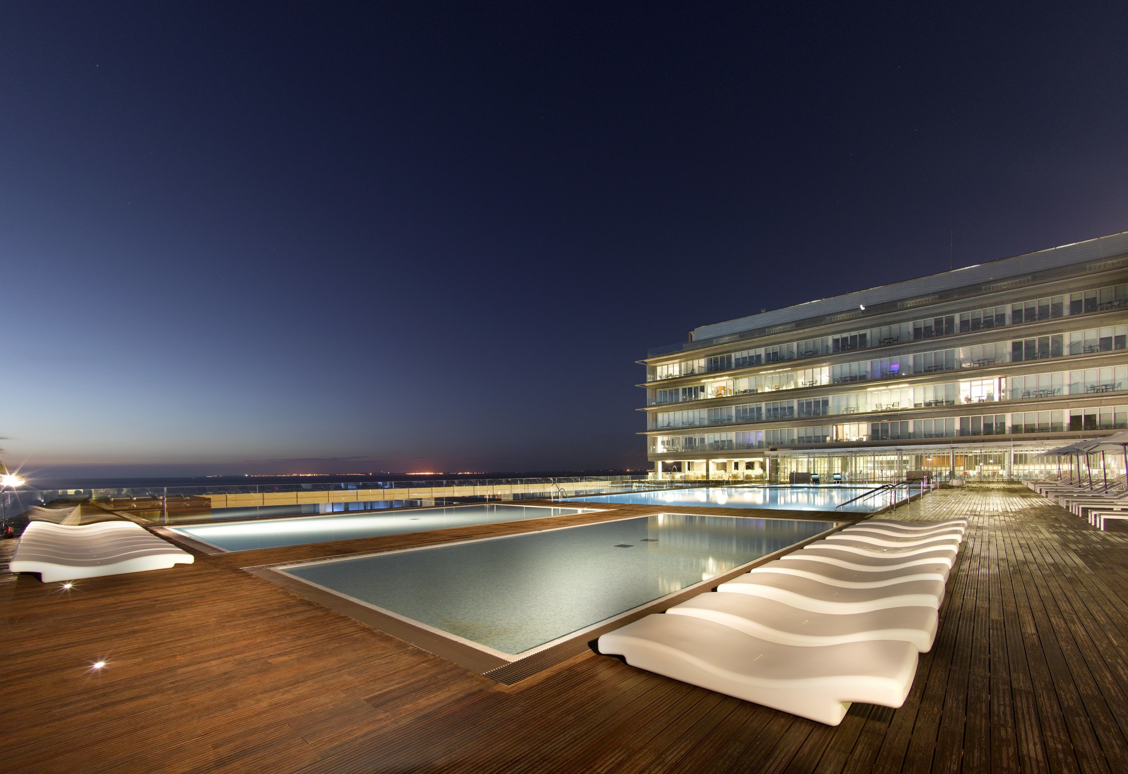 Un espace piscine comprenant deux piscines, des chaises longues et un bâtiment à plusieurs étages doté de balcons.