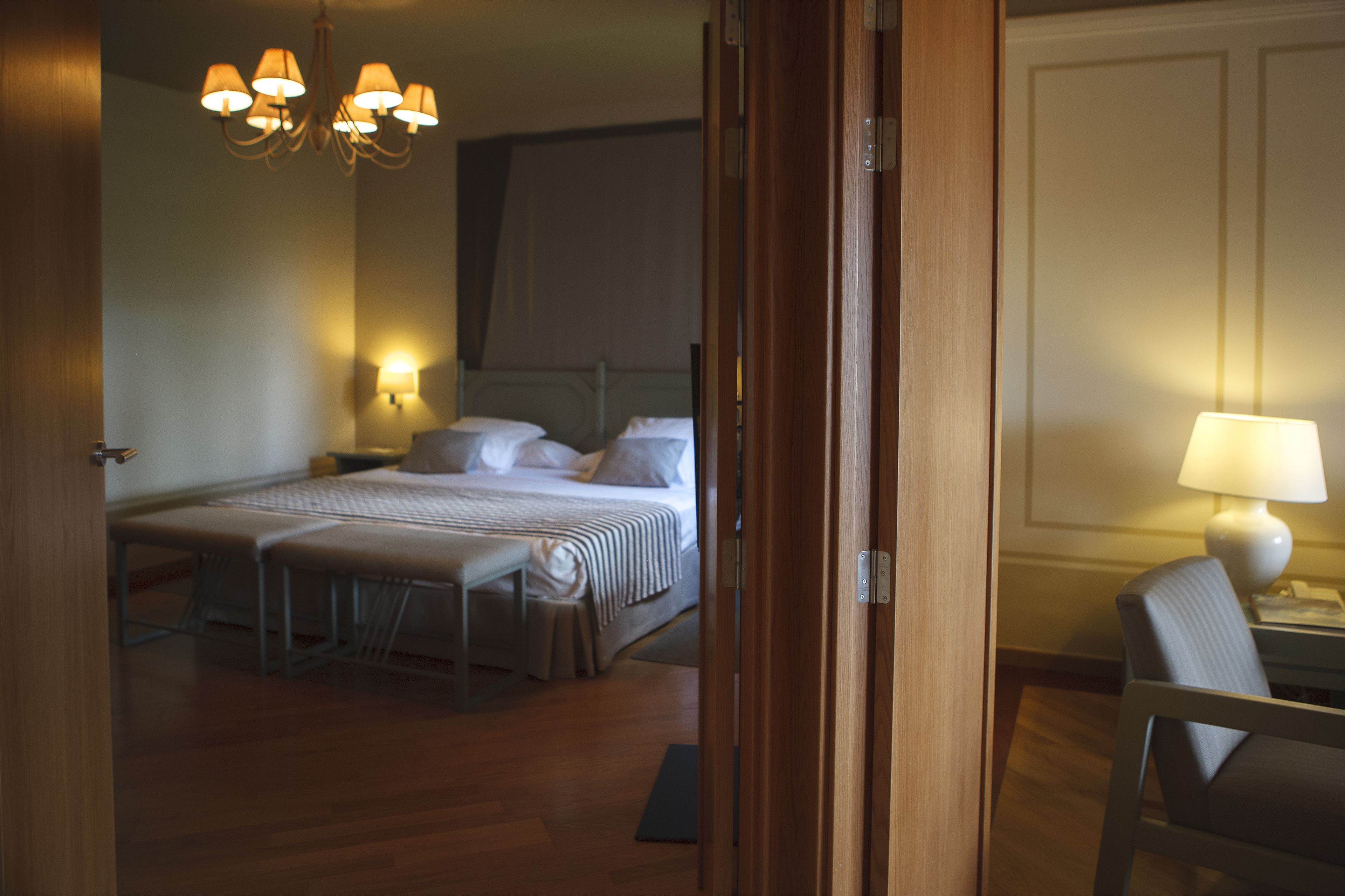 A hotel room with a bed, bedside tables, a chair, a desk, and a chandelier.