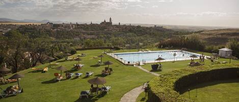 Un área de piscina con sillones reclinables y sombrillas, rodeada de un césped cuidado y una vista a una ciudad lejana con edificios históricos.