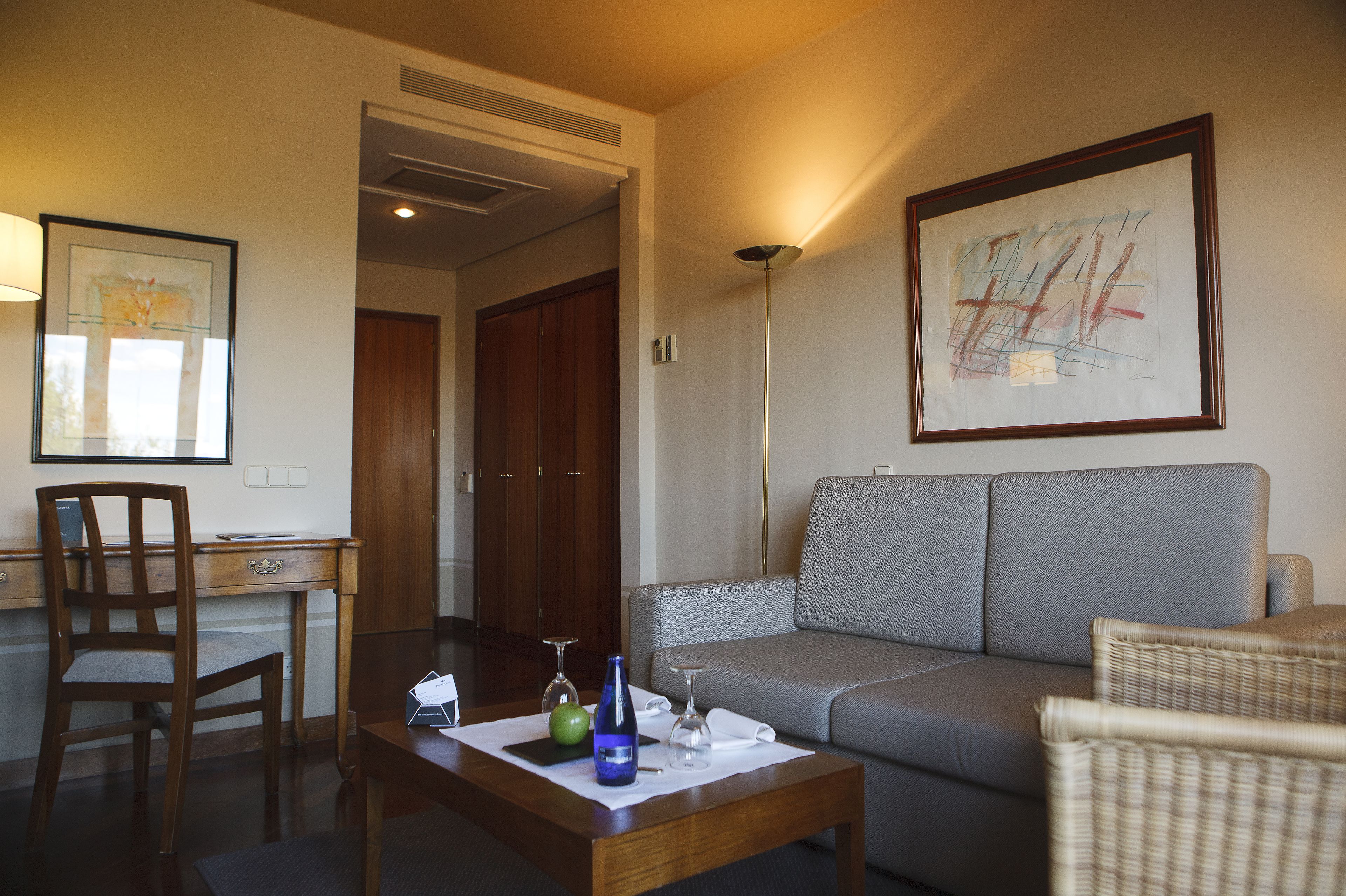 A hotel room with a sofa, a wooden desk, a chair, a coffee table with a vase, and a framed abstract painting.