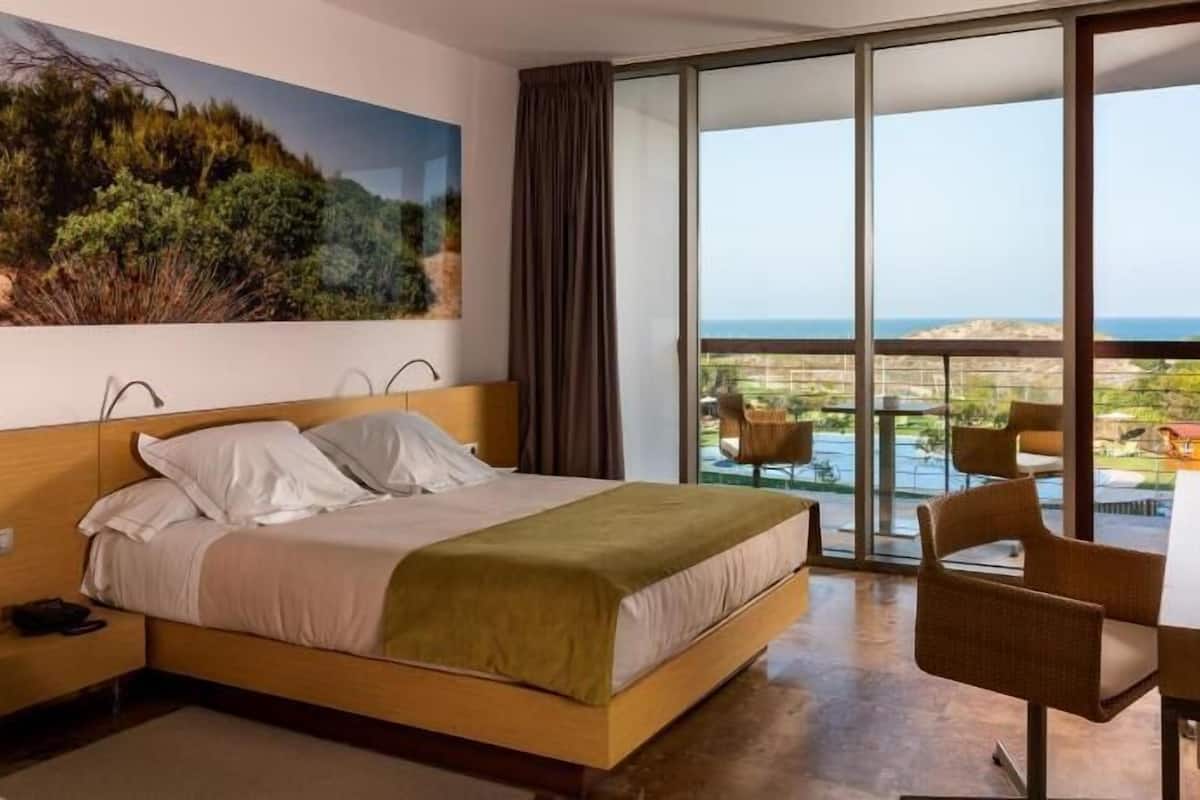 Een hotelkamer met een bed, een bureau, een stoel en een groot raam met uitzicht op de oceaan.