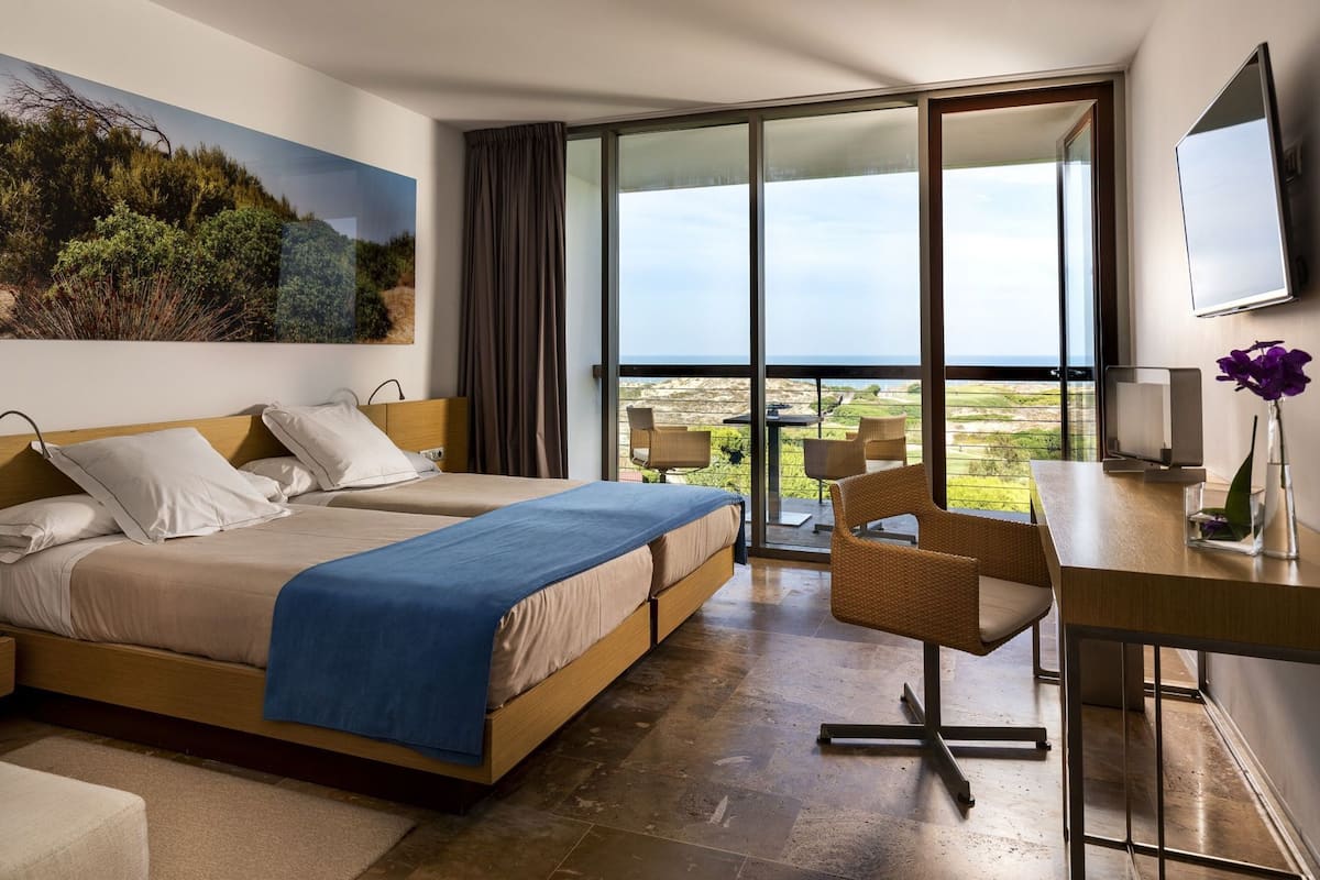 A hotel room with a large bed, a desk with a chair, a television, and a balcony with a view.