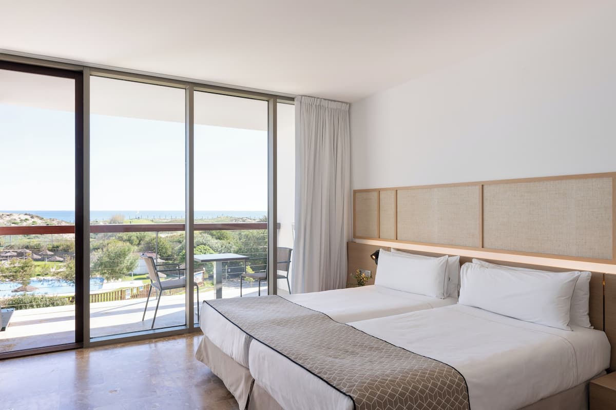 A hotel room with two beds, a large sliding glass door leading to a balcony with a view of a pool and greenery.