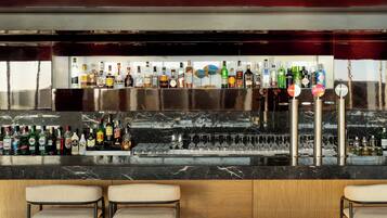 Una zona de bar con mostrador de mármol, varias botellas de licor, vasos y tres taburetes.