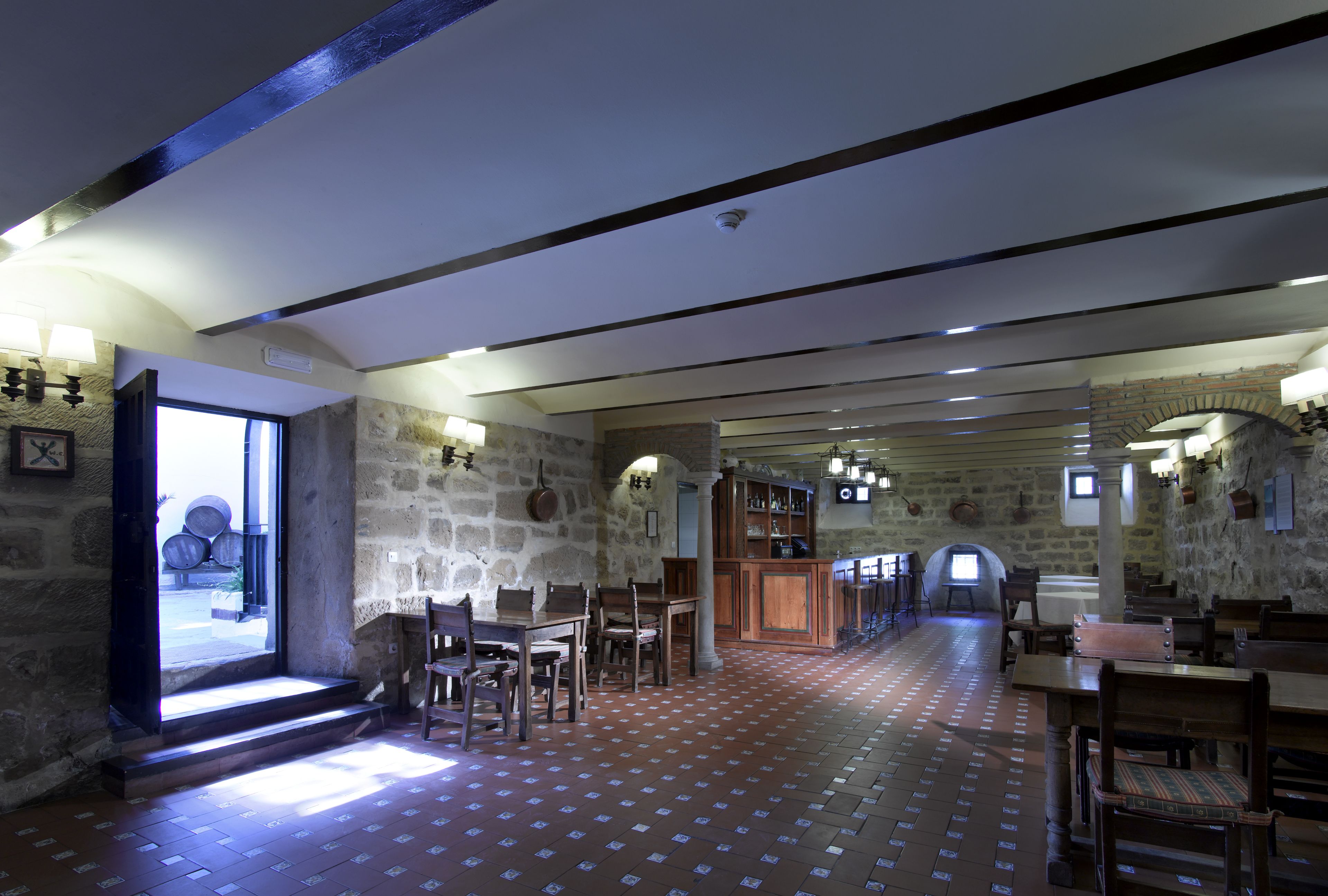 A spacious room with a high ceiling, stone walls, and a long table with chairs. There is a bar area with stools and a television screen.