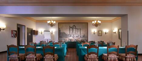 A conference room with tables and chairs, a large painting on the wall, and chandeliers.