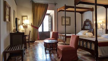 A traditional four-poster bed with a canopy, a desk with a lamp, and a chair in a room with patterned flooring and a large window with curtains.