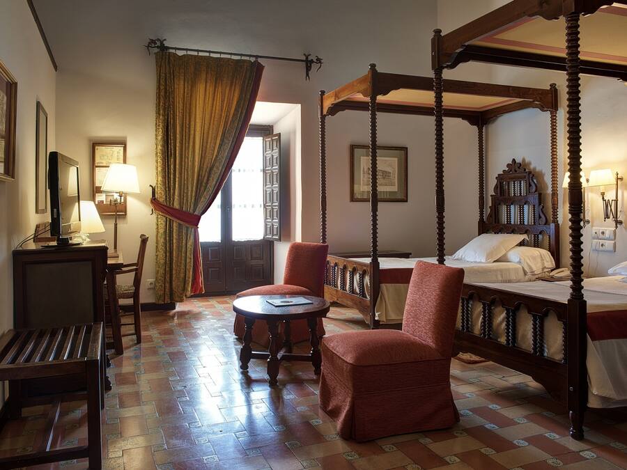 A traditional four-poster bed with a canopy, a desk with a lamp, and a chair in a room with patterned flooring and a large window with curtains.