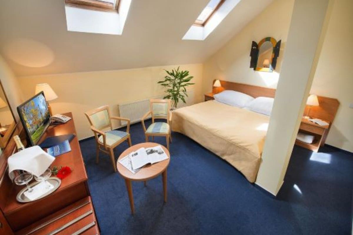 A hotel room with a bed, a desk with a computer, two chairs, a small table, and a plant.