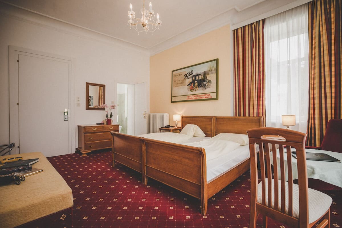 Ein Hotelzimmer mit einem hölzernen Bett, einem Schreibtisch mit Stuhl, einem Schminktisch und einem Spiegel, sowie einem Bild an der Wand.