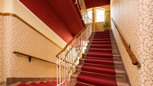 A grand staircase with red carpeted steps and ornate railings, leading to a landing with a potted plant. - Hotel Mozart Bad Gastein (Bad Gastein)