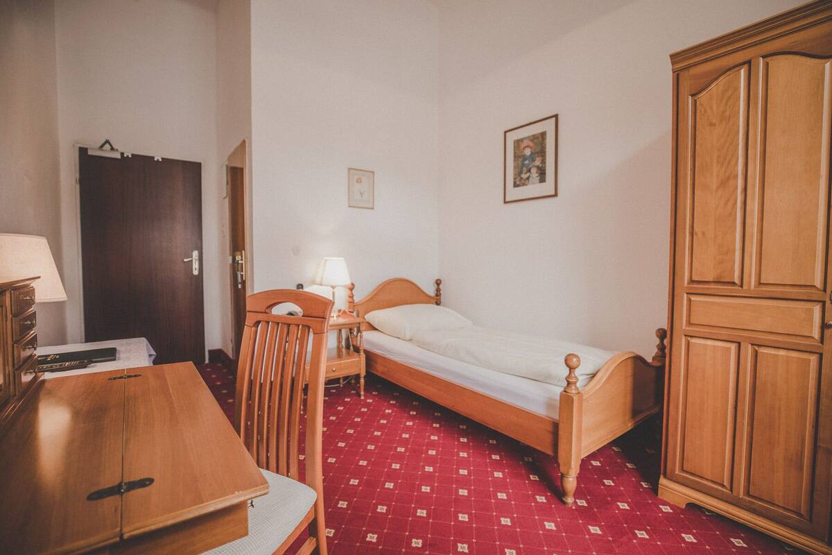 Ein kleines, schlichtes Zimmer mit einem Einzelbett, einem Holzschrank, einem Schreibtisch mit Lampe und einem Stuhl.