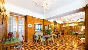 A hotel lobby with a checkered wooden floor, wooden paneling, a dining area with striped chairs, and a chandelier. - Hotel Mozart Bad Gastein (Bad Gastein)