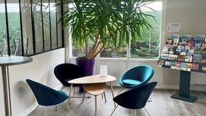Ein moderner Büroraum mit großem Fenster, einer Topfpflanze, zwei blauen Stühlen, einem kleinen Tisch und einer Broschürenauslage.