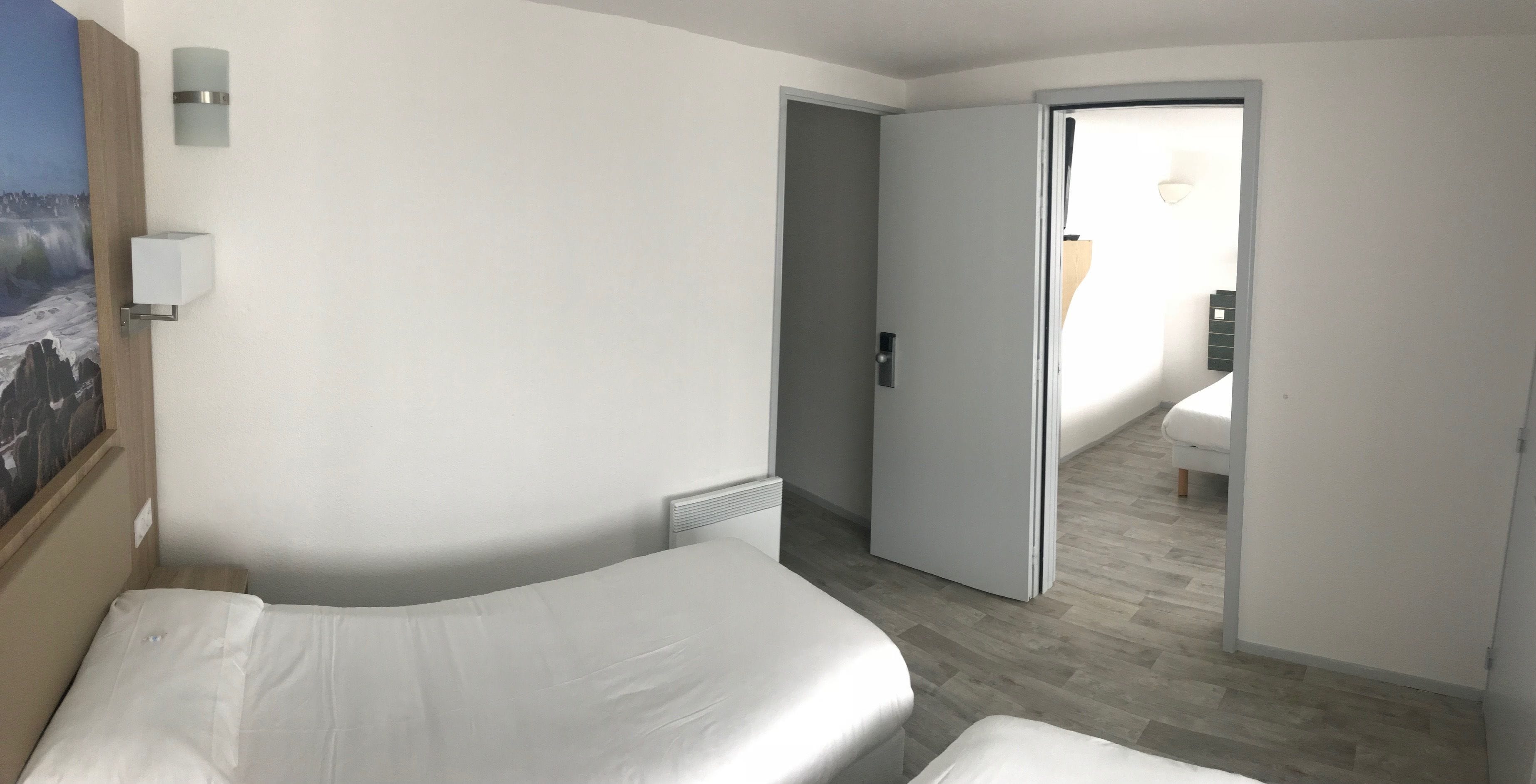 A hotel room with a bed, a nightstand, a wall-mounted light, and a door leading to another room.