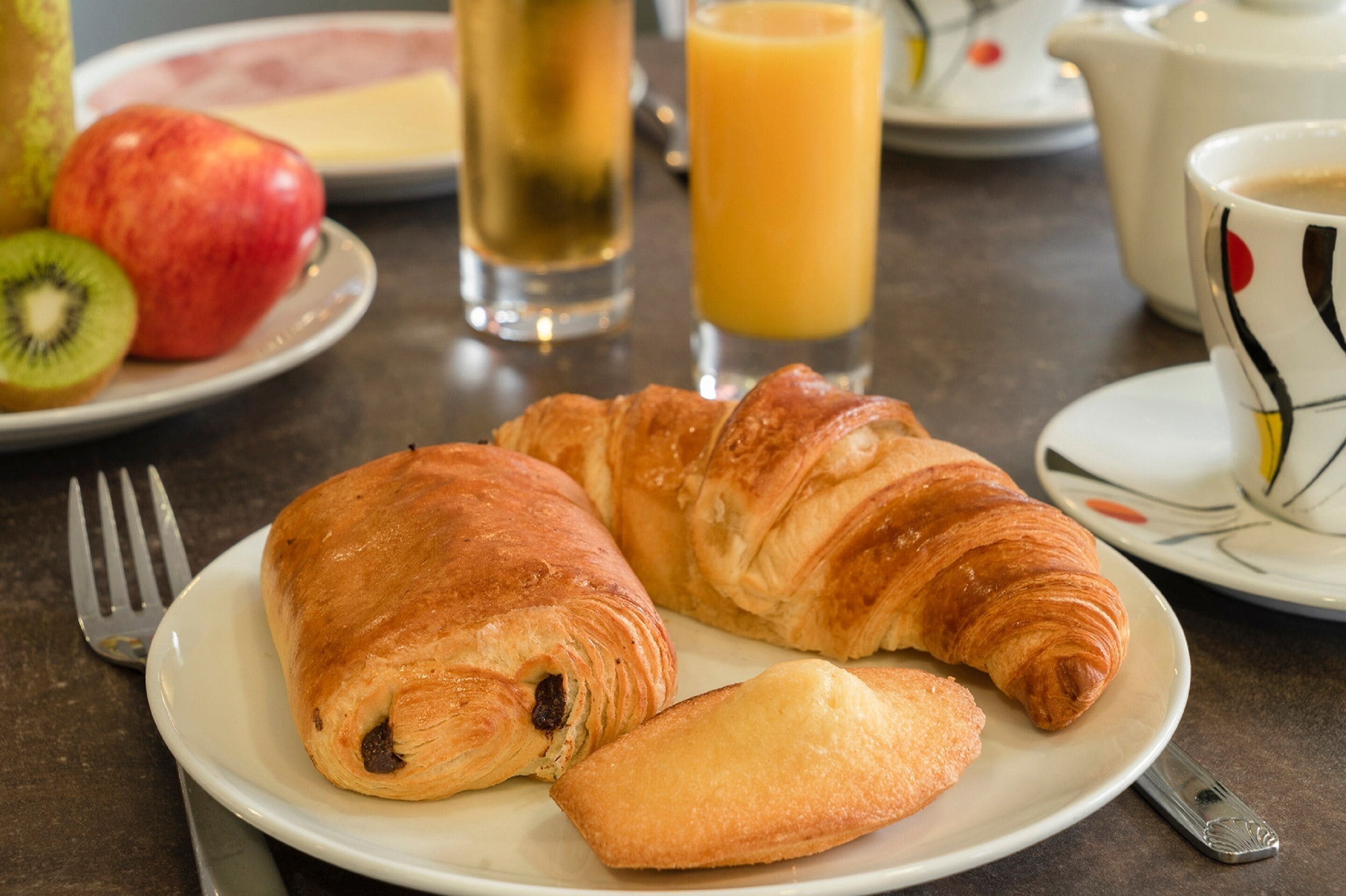 A breakfast setting with croissants, a plate of fruit, a glass of juice, and a cup of coffee.