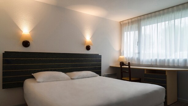 Ein Hotelzimmer mit einem großen Bett, zwei Wandlampen, einem Schreibtisch mit Stuhl und einem Fenster mit leichtem Vorhang.
