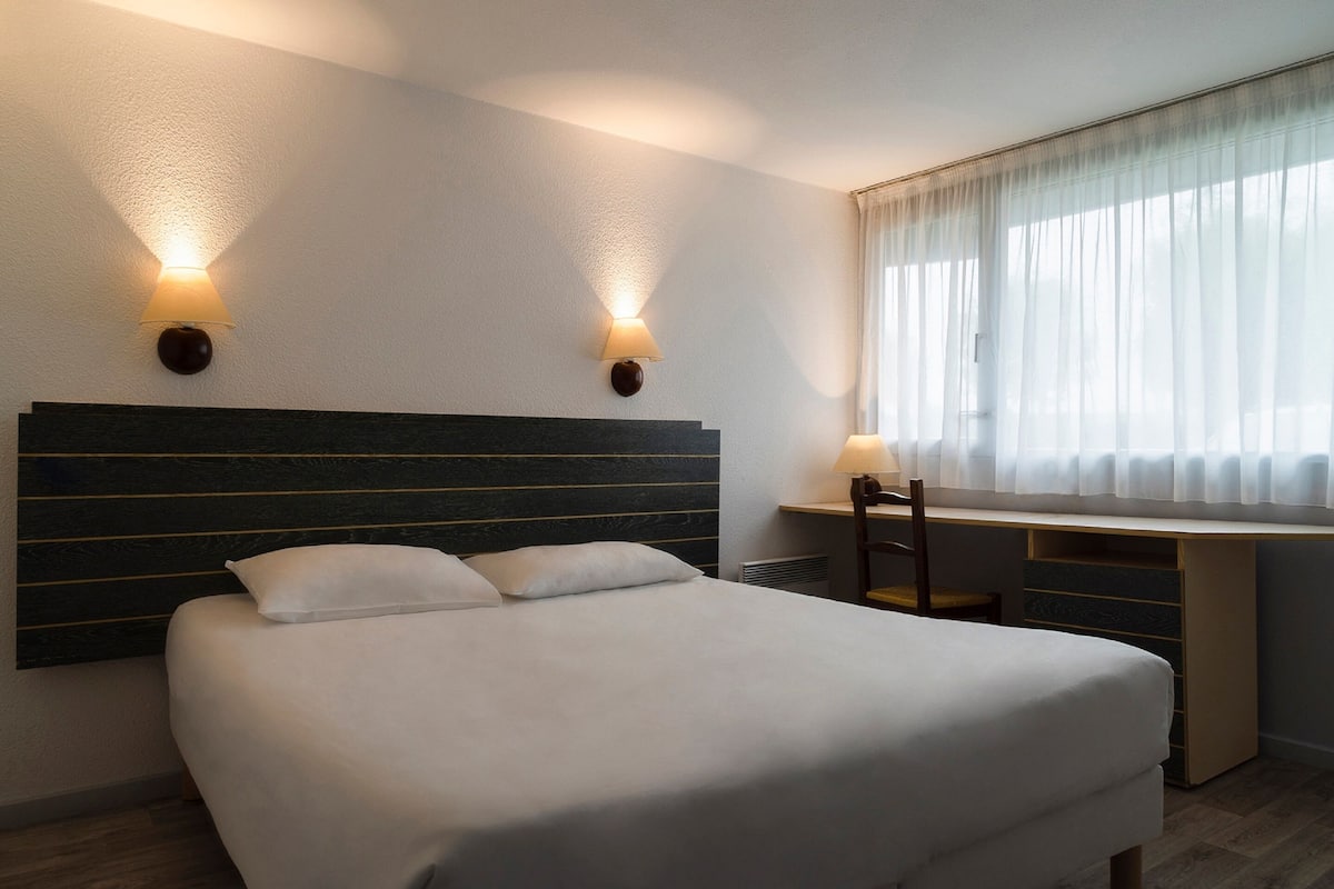 Habitación de hotel con una cama grande, dos lámparas de pared, un escritorio con silla y una ventana con cortinas translúcidas.