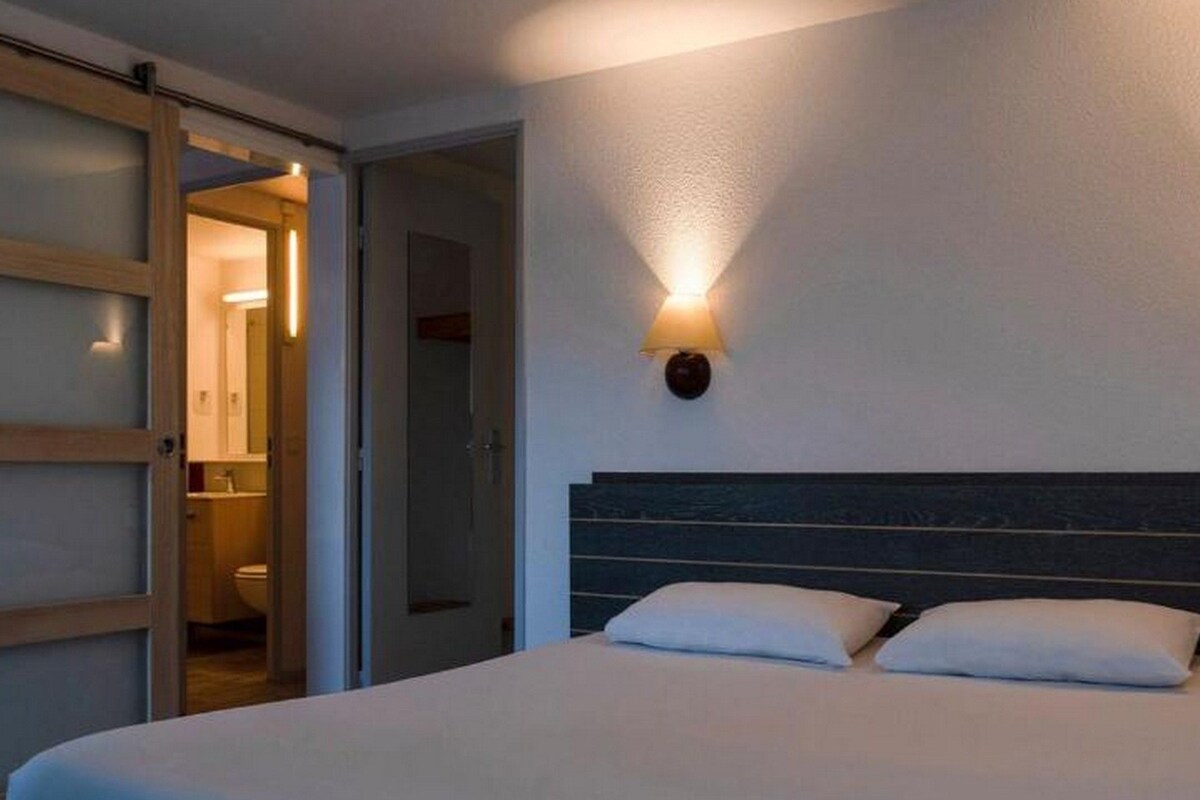 Habitación de hotel con cama, mesita de noche, lámpara de pared y puerta abierta que da a un baño.