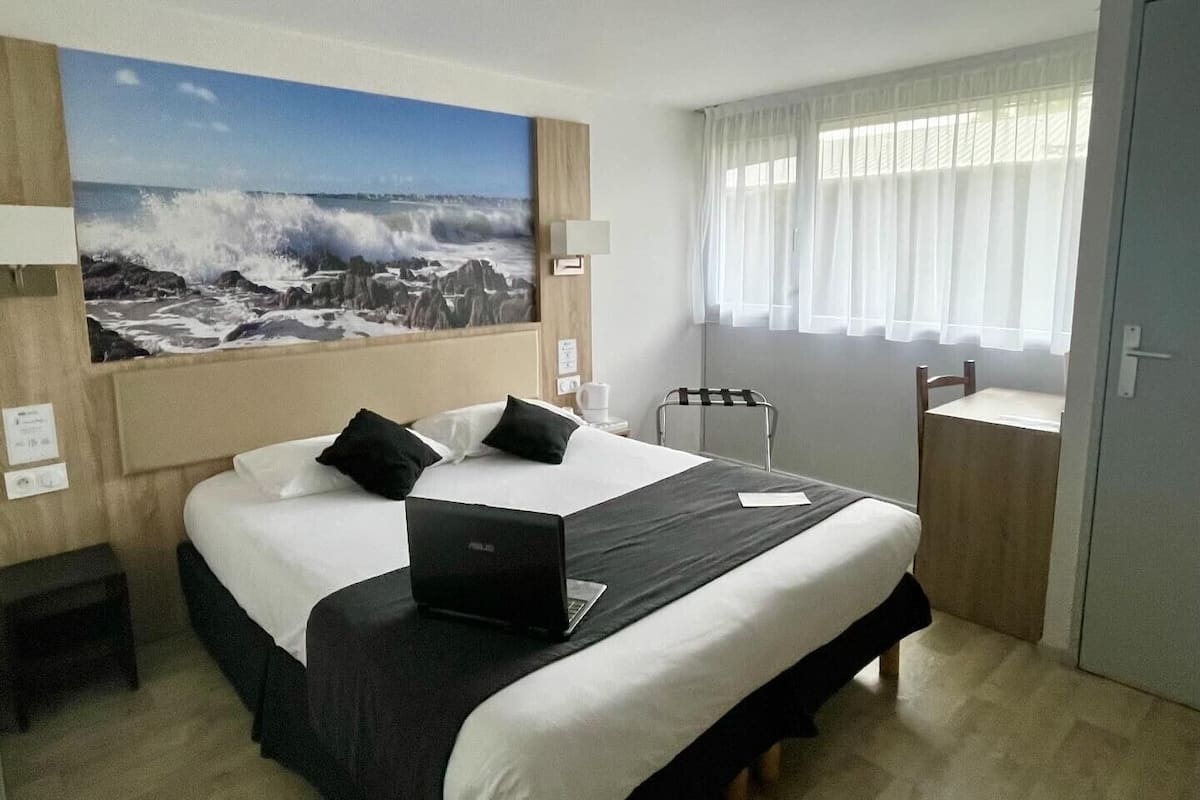 Habitación de hotel con una cama grande, una laptop sobre la cama, un cuadro de un paisaje marino en la pared, una ventana con cortinas translúcidas y una pequeña mesa con una silla.