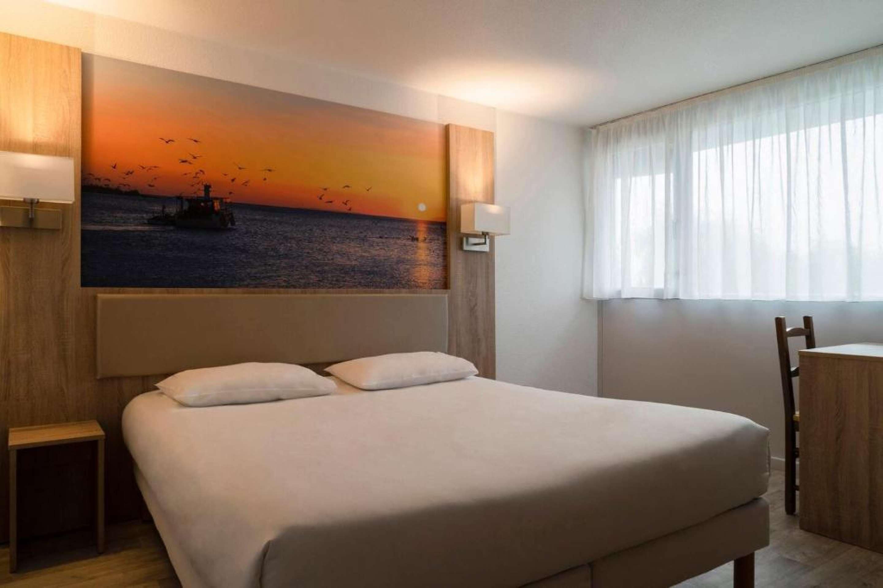 Een hotelkamer met een groot bed, een kleintje tafeltje, een stoel, een schilderij van een zonsondergang boven de oceaan en een raam met dunne gordijnen.