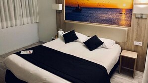 Ein Hotelzimmer mit Bett, Nachttisch, Lampe und einem Gemälde, das einen Sonnenuntergang über dem Meer zeigt.
