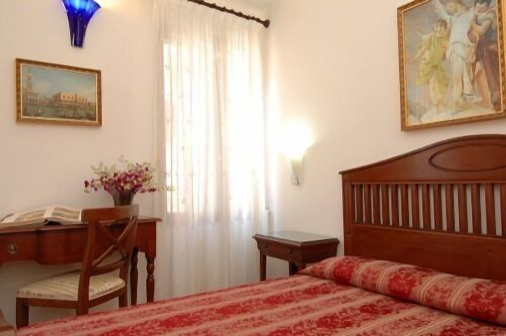 Un dormitorio con una cama de madera, un escritorio con una silla, un jarrón con flores y dos cuadros en las paredes.