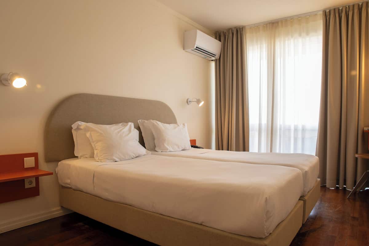 A hotel room with a double bed, a nightstand, an air conditioning unit, and a window with curtains.