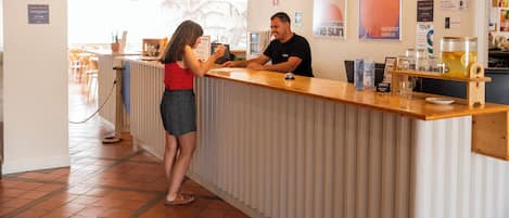 Uma área de recepção com balcão de madeira, uma mulher e um homem atrás dele, e diversos serviços como uma máquina de café e copos.