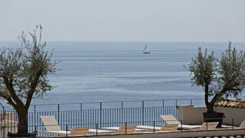 A seaside view with a sailboat on the water, lounge chairs, and a railing.