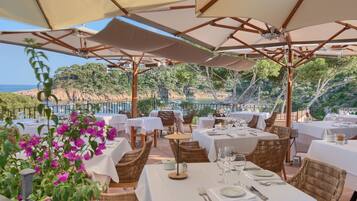 An outdoor dining area with tables and chairs, umbrellas, and a view of the sea.