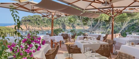 An outdoor dining area with tables and chairs, umbrellas, and a view of the sea.