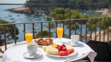 Una mesa de desayuno con cruasanes, fruta, café y jugo, con vistas a la costa donde se ven barcos y vegetación.