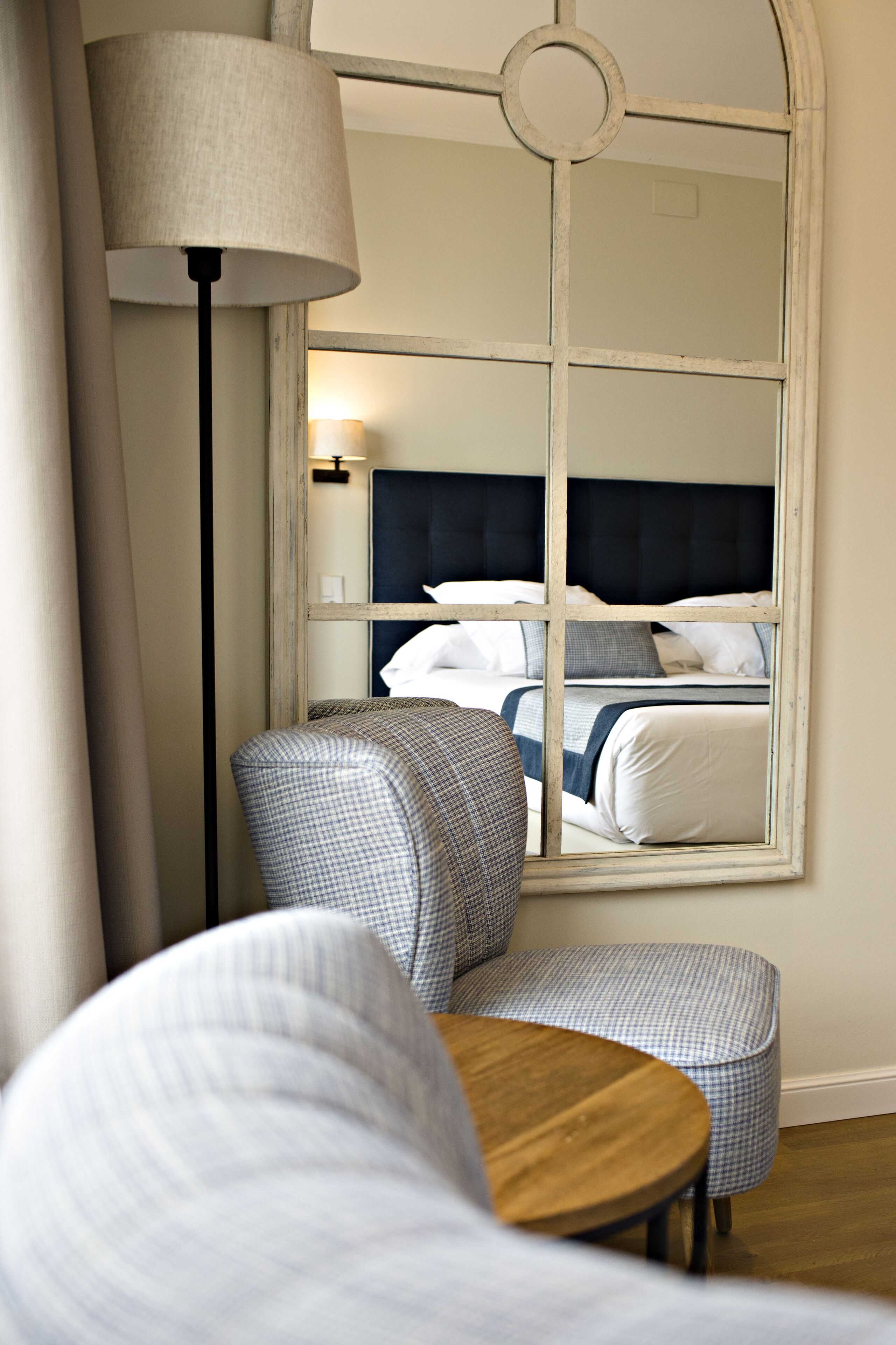 A hotel room with a large mirror reflecting a bed, a lamp, and a checkered chair.