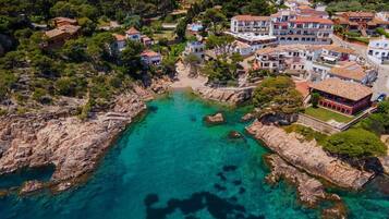 Vista aérea de un pueblo costero con edificios, aguas turquesas cristalinas, costa rocosa y abundante vegetación.