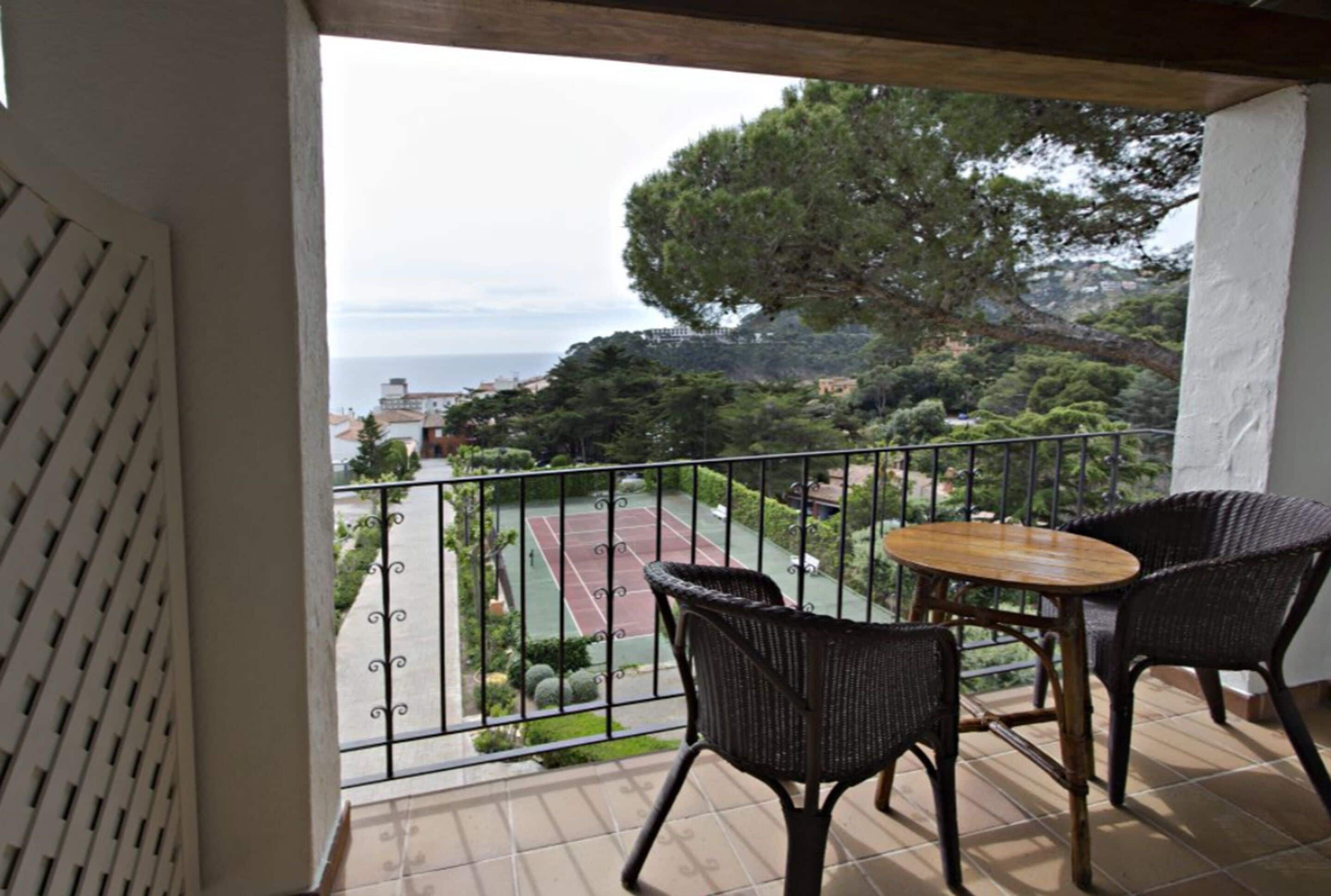 A balcony with wicker chairs and a table, overlooking a tennis court and a scenic coastal view.