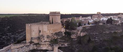 Un castillo de piedra histórico con una torre destacada, rodeado por una muralla fortificada y situado en una colina con vistas a un pueblo.