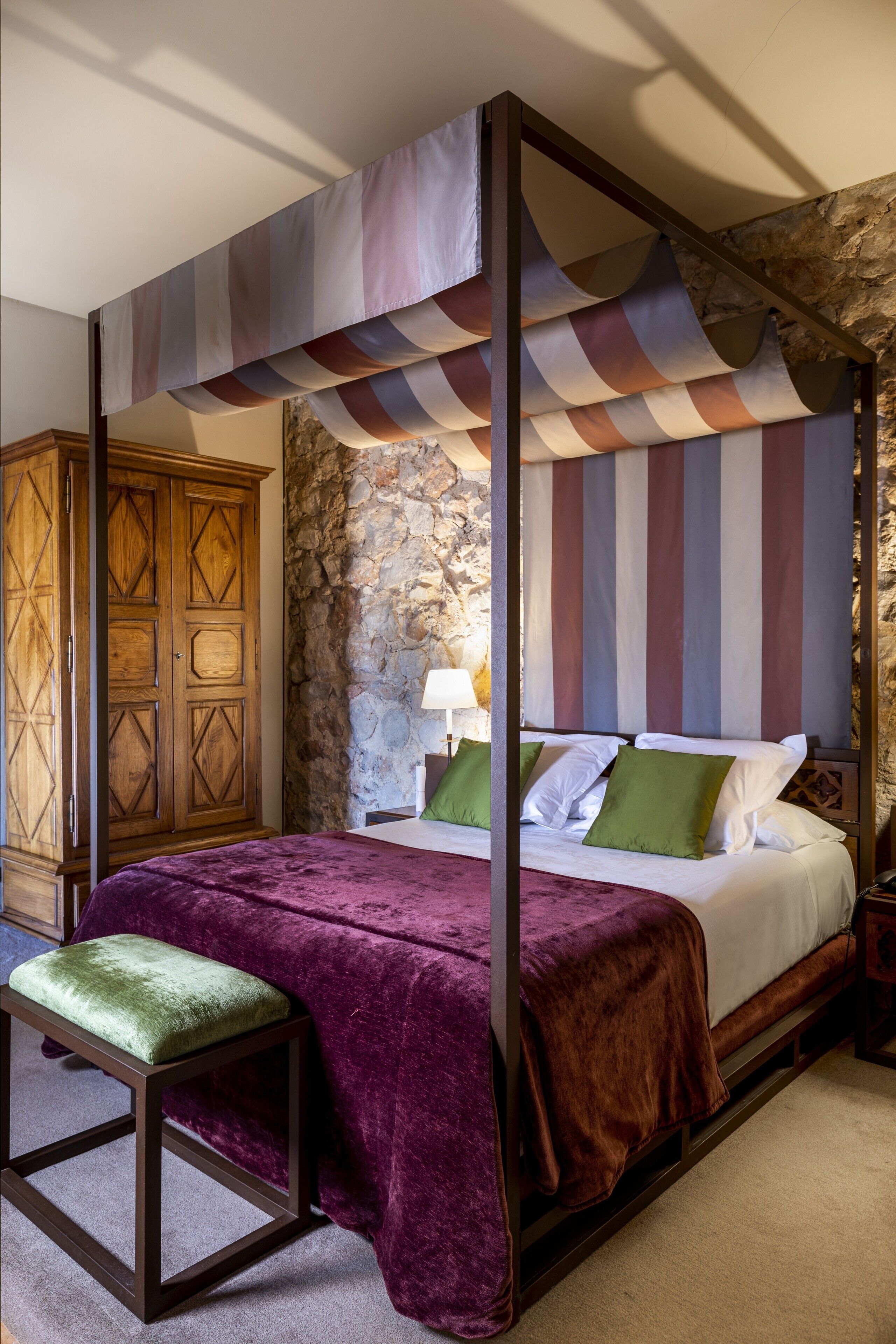 A four-poster bed with a striped canopy, a stone wall, and a wooden wardrobe.